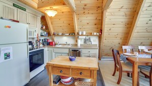 A cozy kitchen with wooden cabinets, a refrigerator, microwave, and a dining area with a table and chairs. - Upper Peninsula Lakefront A-frame Cabin w/ Dock (Channing)