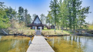A wooden dock leading to a cabin with a glass front, surrounded by trees and a body of water. - Upper Peninsula Lakefront A-frame Cabin w/ Dock (Channing)