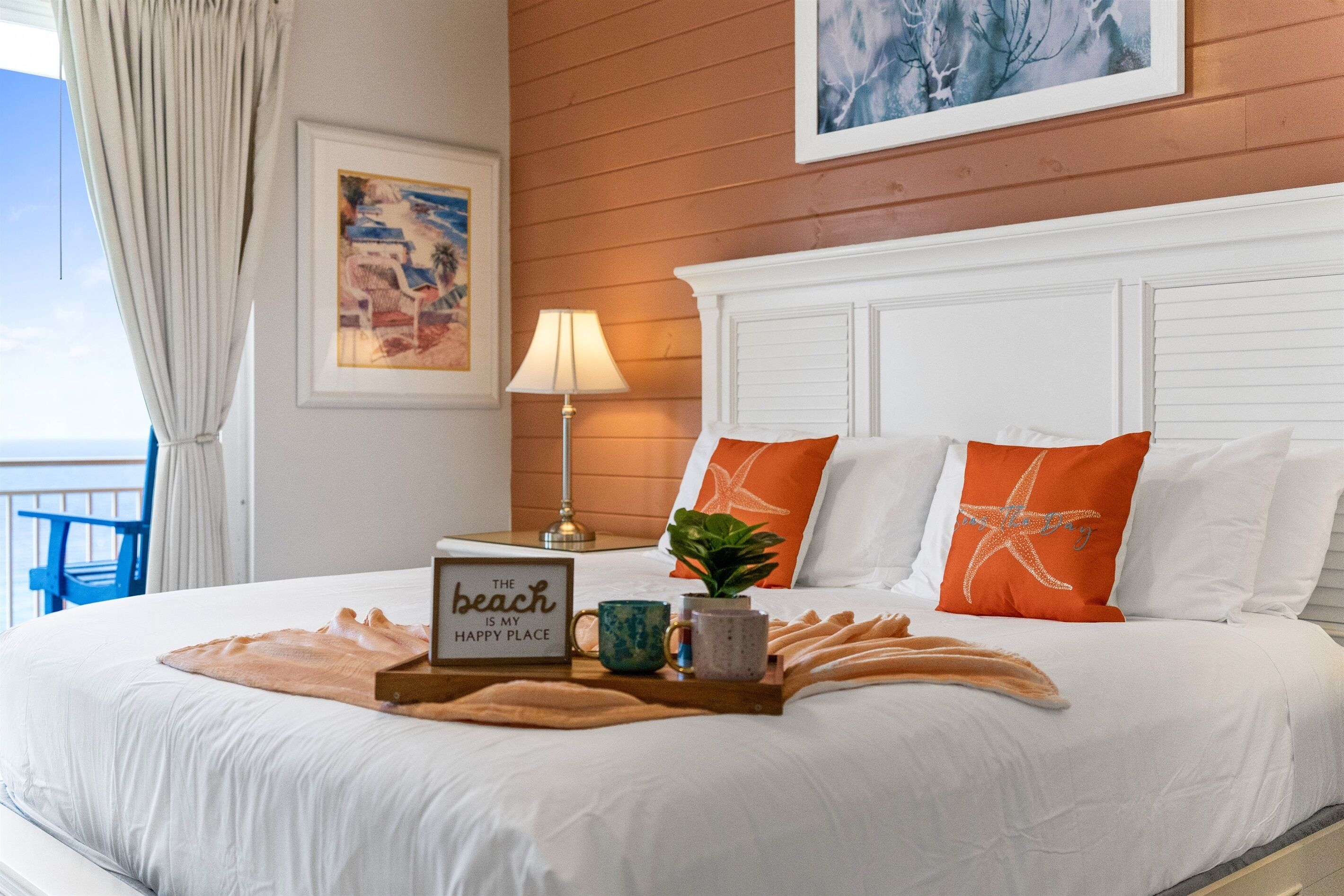 Een netjes opgemaakt bed met een strandkussen, een kleine plant en een dienblad met een strandbord en twee mokken.