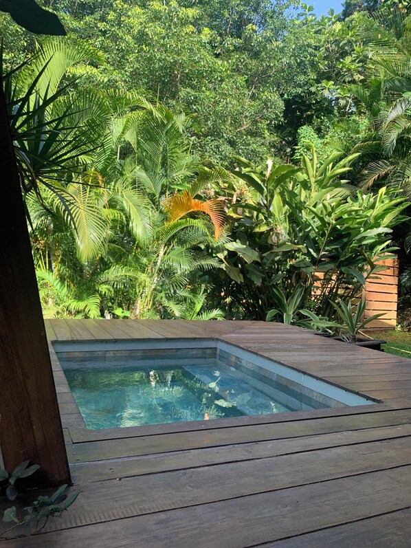 A rectangular hot tub surrounded by lush greenery and a wooden deck. - Cottage in the heart of the rainforest (Sainte-Rose)