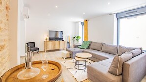 A modern living room with a grey sofa, a wooden coffee table, a TV on a wooden cabinet, and a large window with curtains.