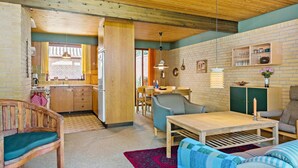 Ein gemütlicher, gut beleuchteter Raum mit Holzbalken an der Decke, einem Essbereich mit Tisch und Stühlen, einer Küche mit Kühlschrank und einem Wohnbereich mit Sofa und Couchtisch.