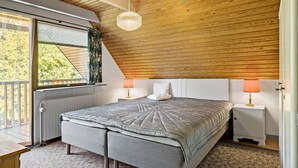 Ein Schlafzimmer mit schrägen Wänden, einem Bett mit grauer Decke, zwei Nachttischlampen und einem Fenster mit Vorhängen.