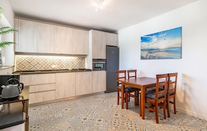 A compact kitchen with wooden cabinets, a built-in oven, and a refrigerator. There's a dining table with four chairs and a patterned floor.
