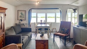 A cozy living room with a sofa, a wooden coffee table, and a dining table with chairs. There is a large window providing a view of the outdoors. - Stunning (sta)caravan in Reeuwijk (Reeuwijk)