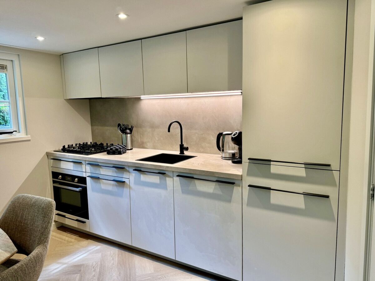 Eine moderne Küche mit Einbauherd, Spüle und Kühlschrank. Über der Spüle befindet sich ein Fenster und im Vordergrund ein Stuhl.