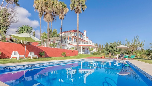 A swimming pool with a clear blue water, surrounded by palm trees and a large house with a red wall and white balcony. - Gorgeous apartment in Malaga with house sea view (Malaga)