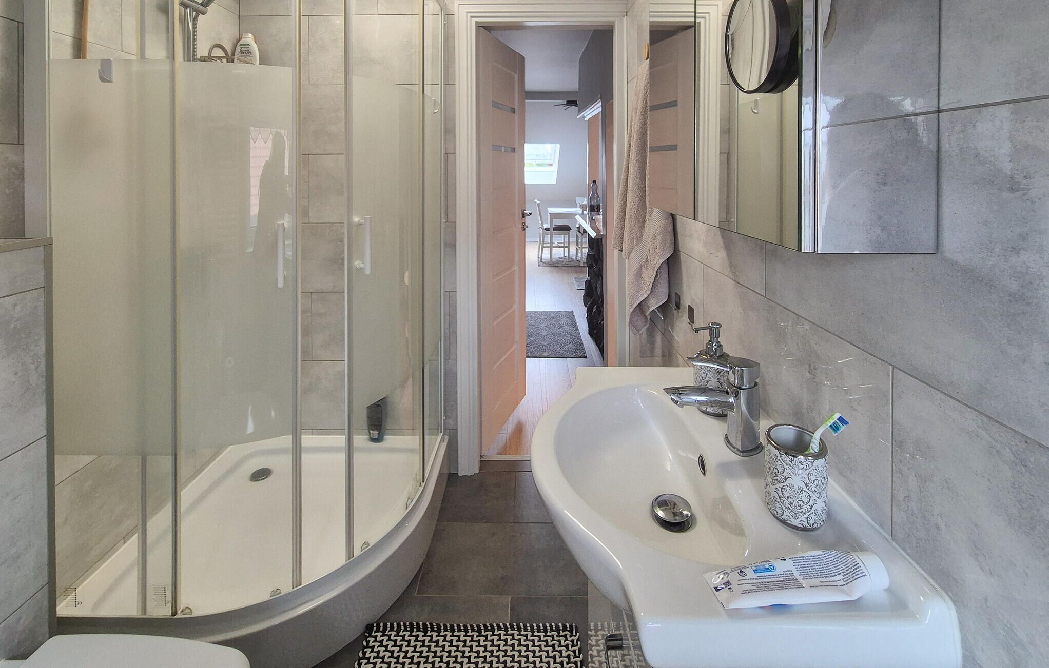 Ein modernes Badezimmer mit einer Glaskabine zur Dusche, einem weißen Waschbecken inklusive Spiegel und einem gefliesten Boden.