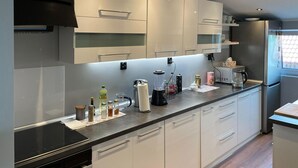 Una cocina moderna con armarios blancos, una placa de inducción negra y un horno empotrado. Hay varios electrodomésticos y utensilios en la encimera, incluyendo una licuadora, un hervidor y una lata de refresco.