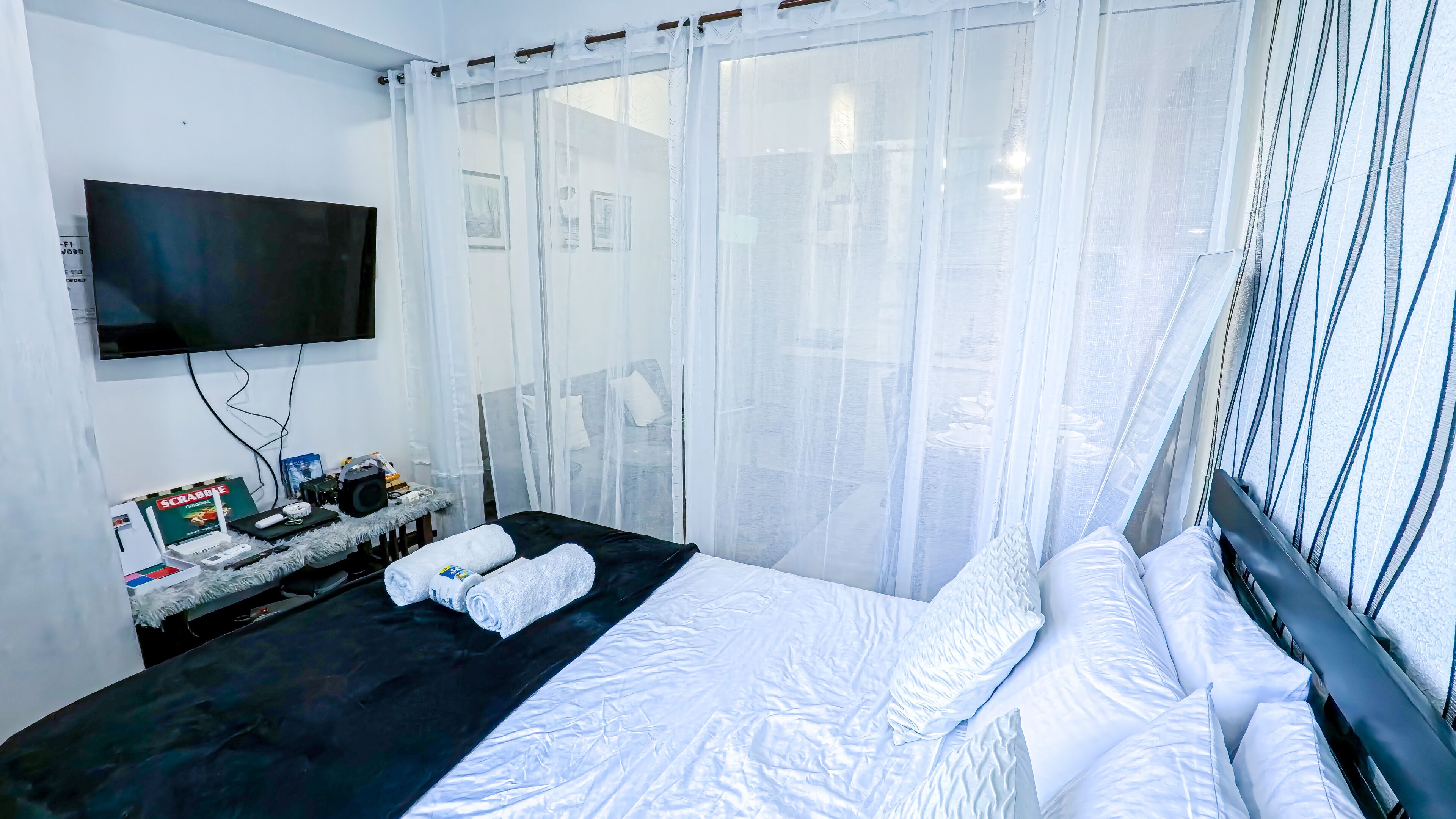A hotel room with a bed, a flat-screen TV mounted on the wall, a small table with a Scrabble game and other items, and sheer curtains on the windows.