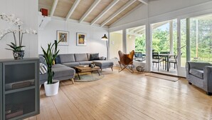Een ruime woonkamer met een houten plafond, een grijze bank, een salontafel en een lederen fauteuil. Grote ramen bieden uitzicht op het omliggende landschap.