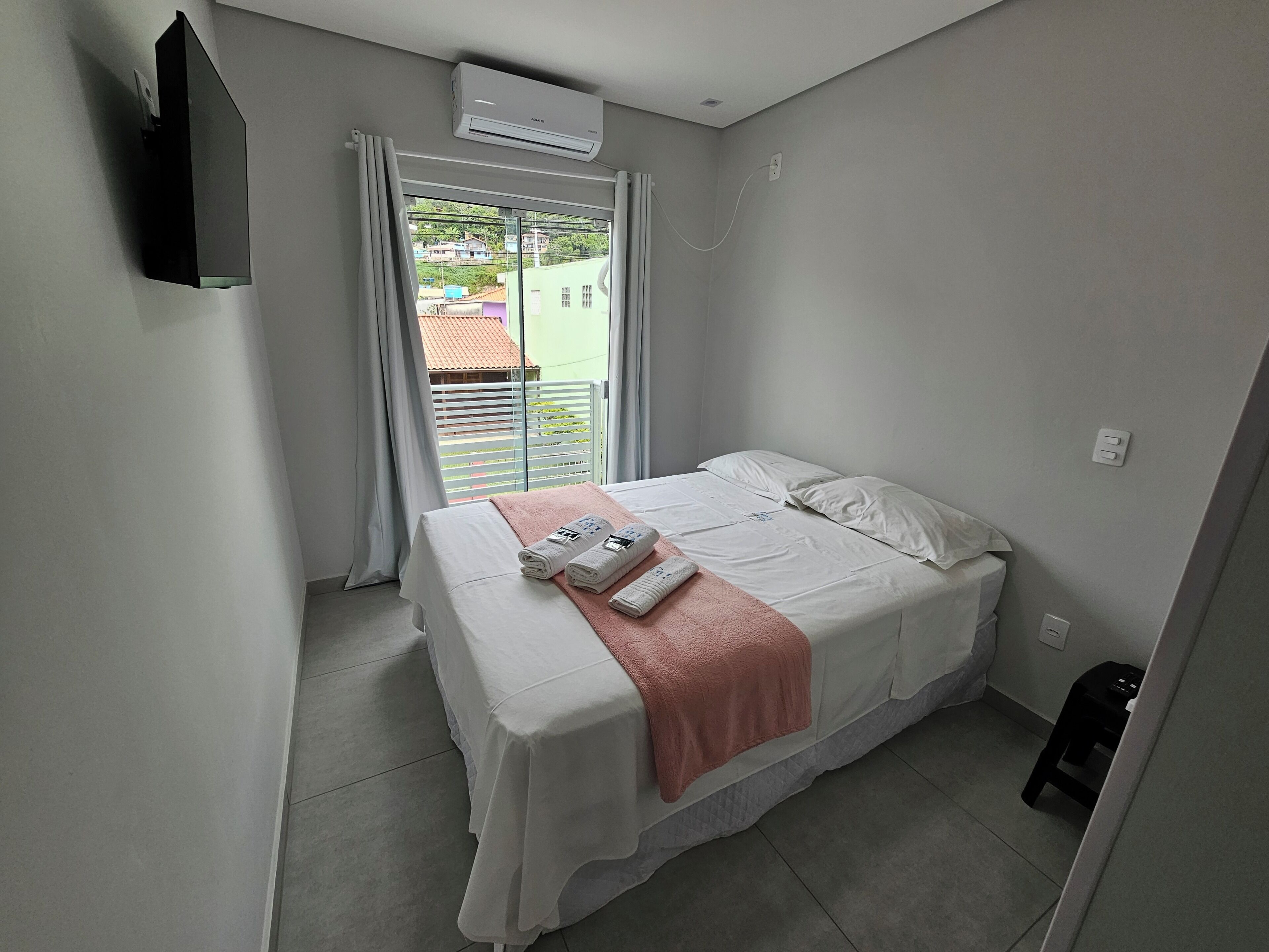 A bedroom with a bed, a window with curtains, an air conditioning unit, and a small stool.