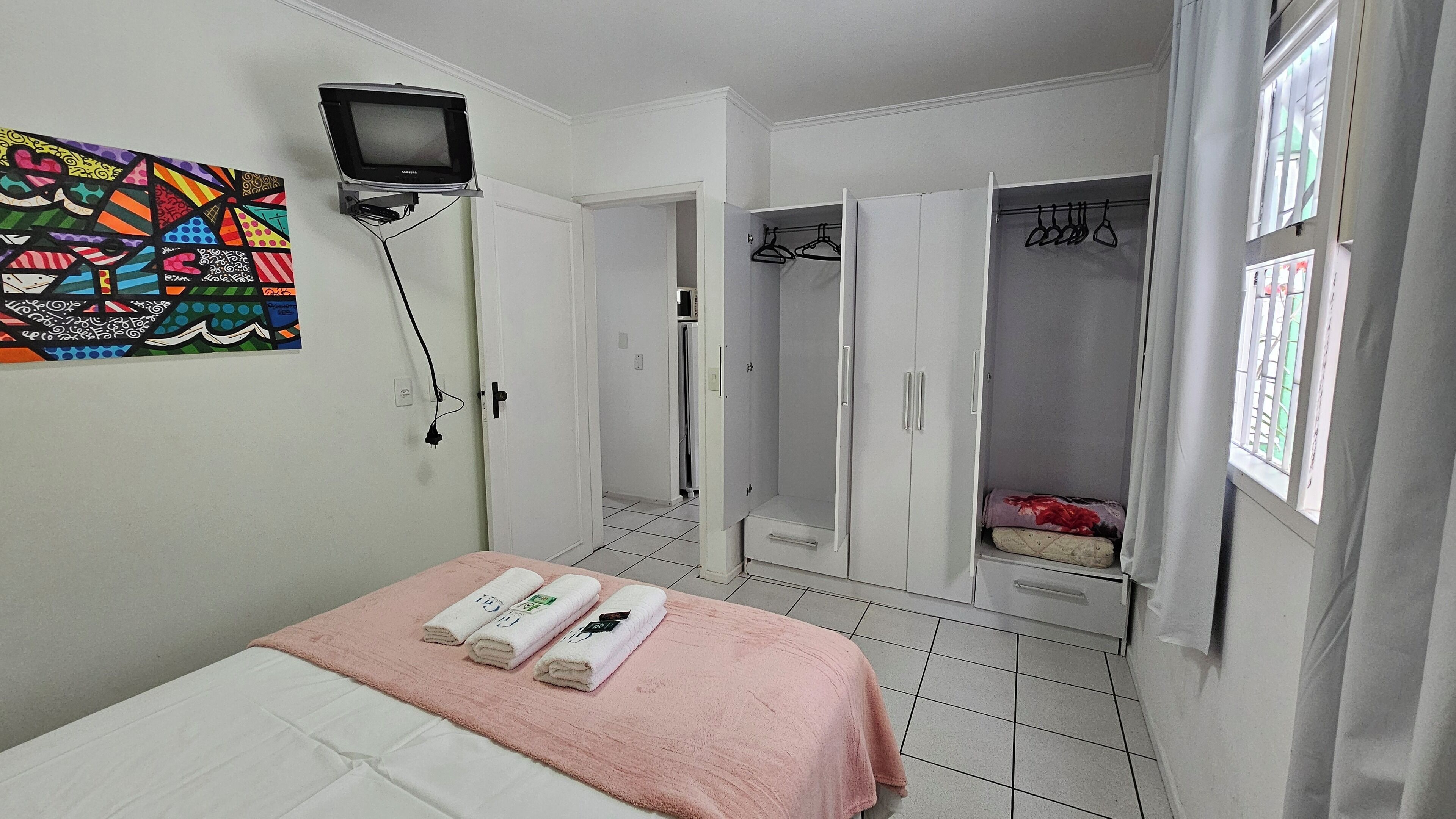 Una camera da letto con un letto, una televisione a muro, un armadio pieno di vestiti e una finestra con tende.