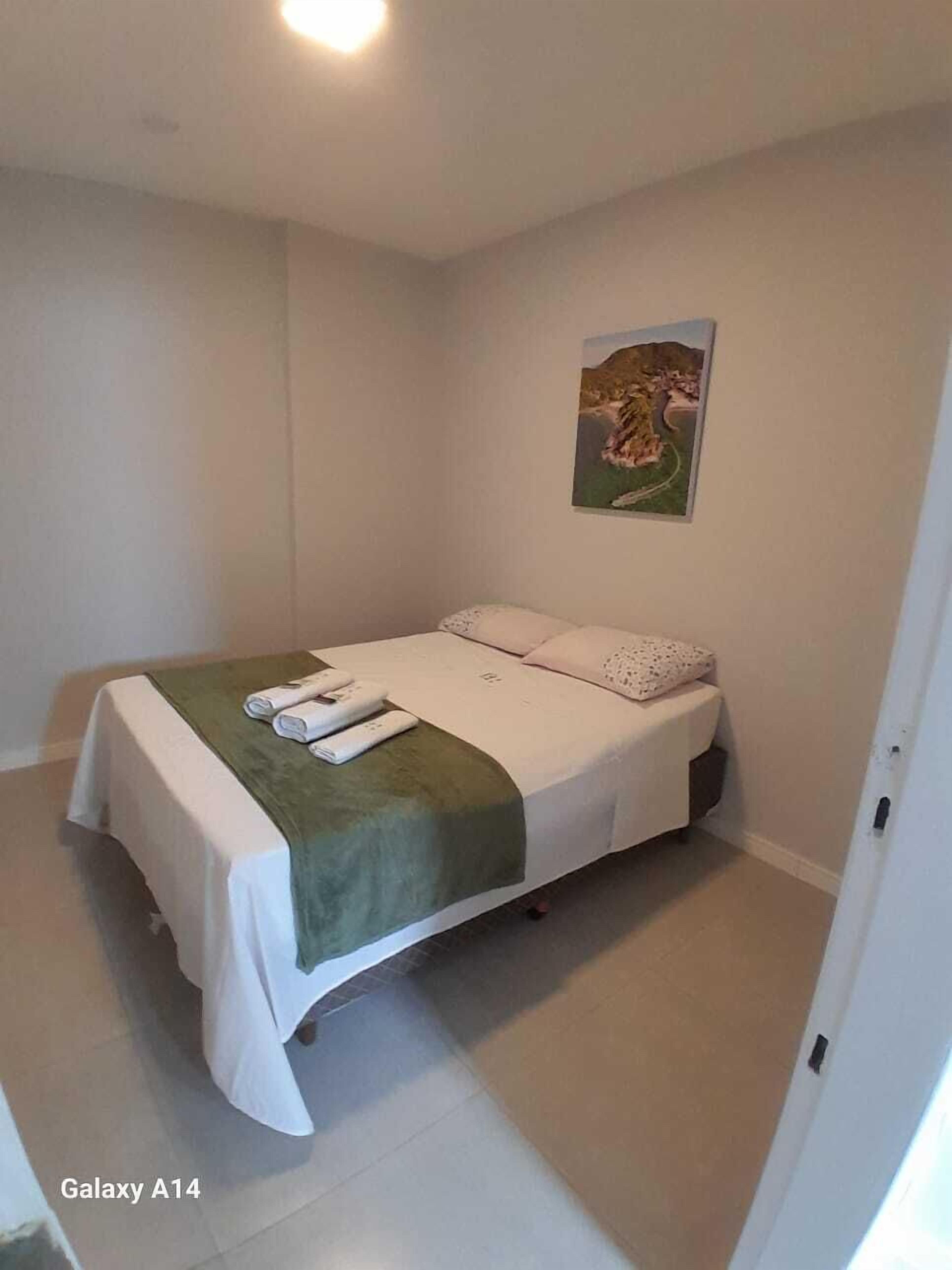 Una cama individual con una manta verde y ropa de cama blanca, una mesita de noche pequeña y un cuadro colgado en la pared.