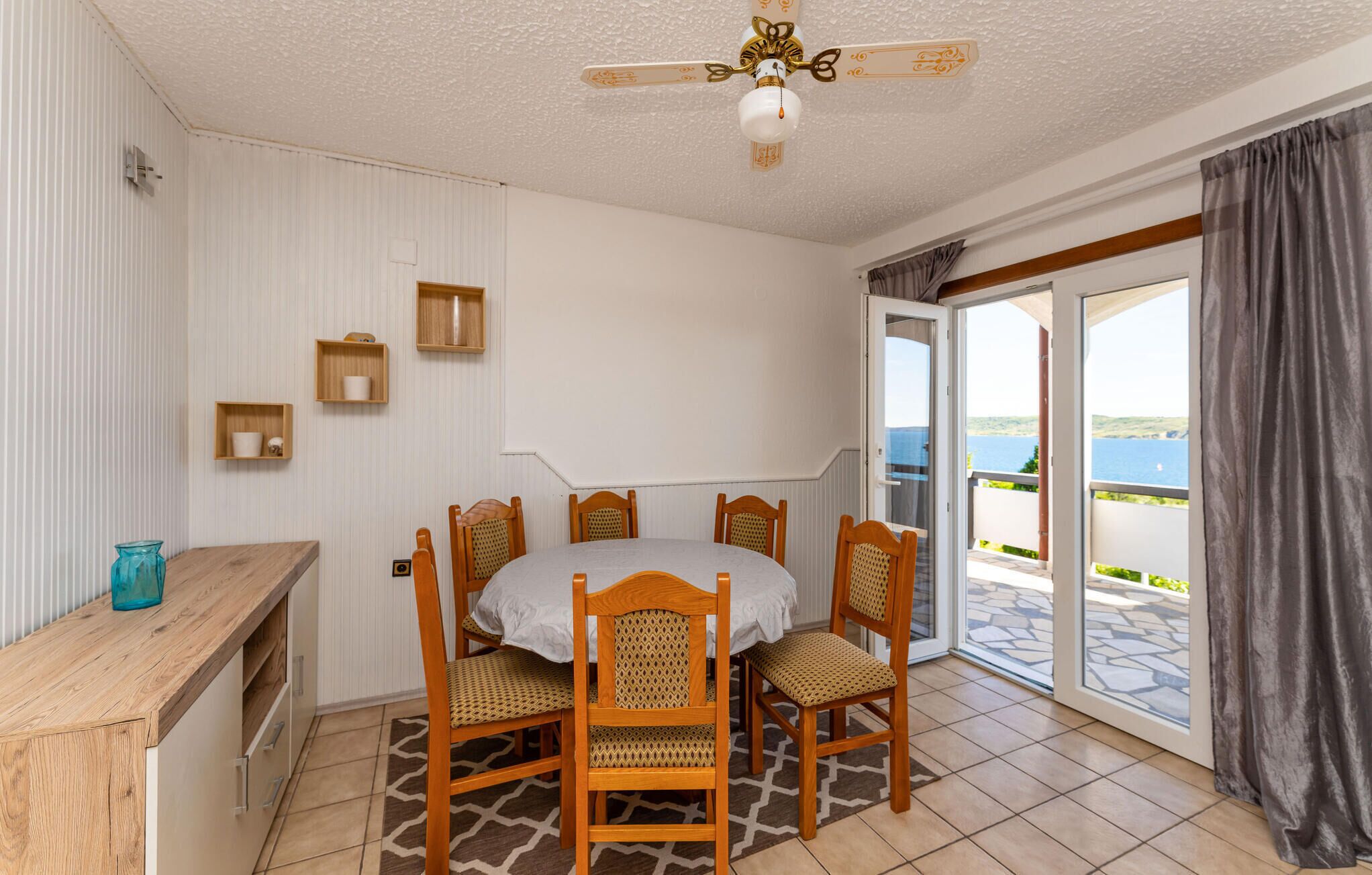 A dining area with a table and chairs, a ceiling fan, and a view of the sea through a glass door.