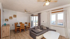 Ett vardagsrum med en soffa, ett soffbord, en matplats med stolar och ett bord, en takfläkt och utsikt över havet genom fönstren.