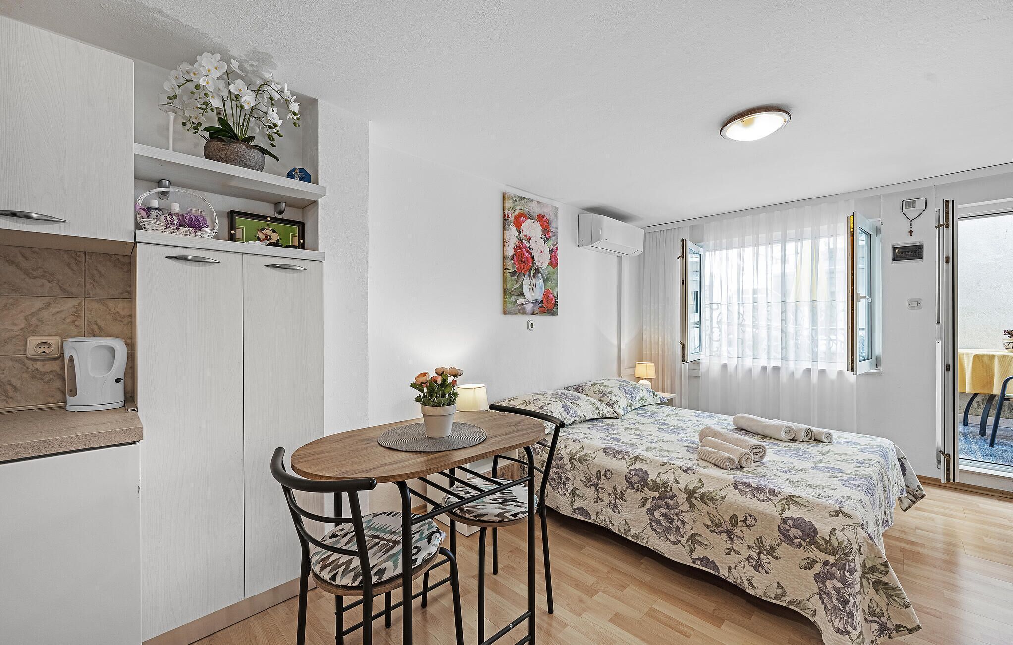 Quarto com cama, mesa de cabeceira, cadeira, janela com cortinas, um quadro na parede e um espaço de kitchenette com cafeteira.