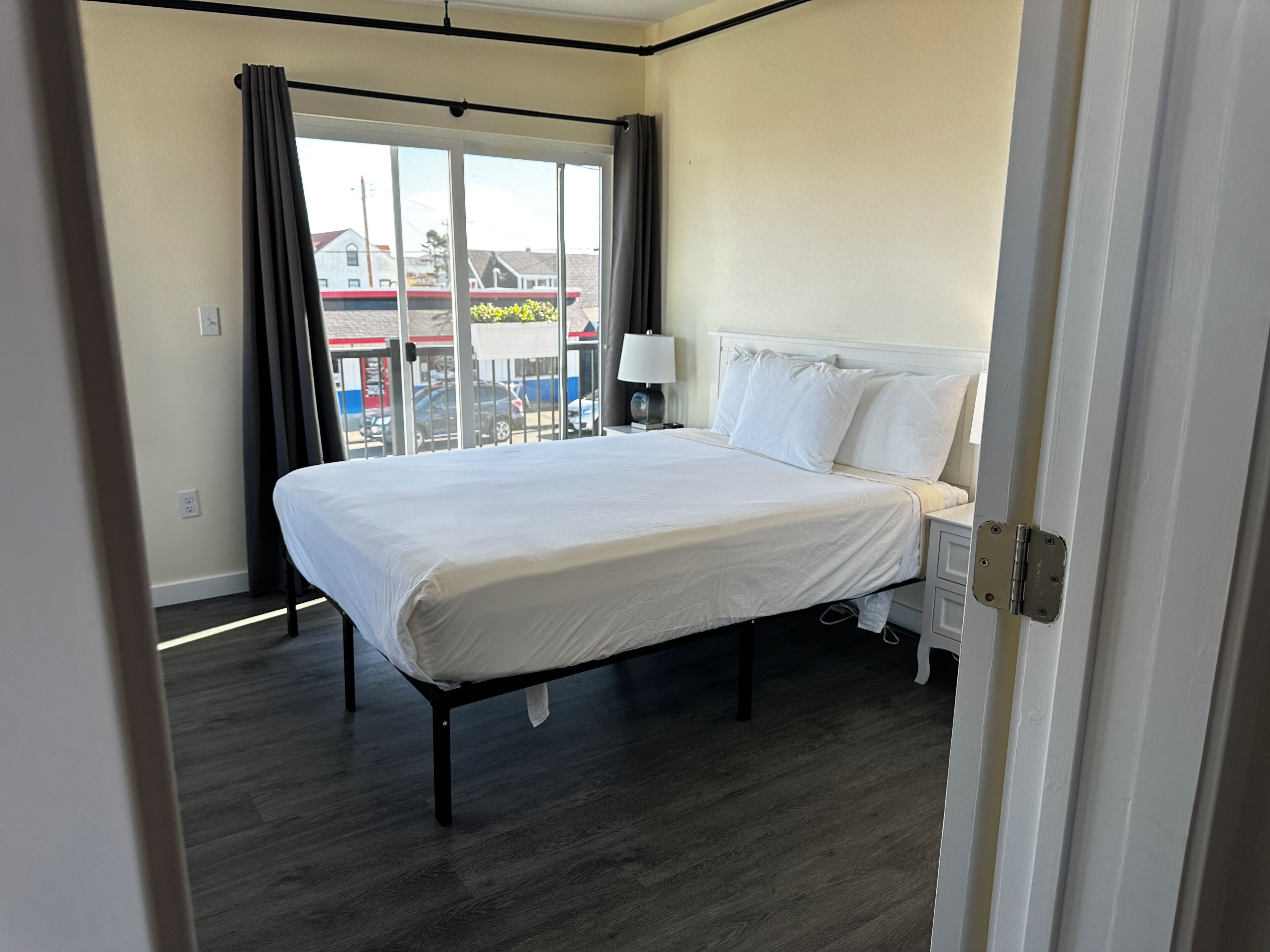 A hotel room with a bed, a nightstand, a lamp, and a view of the outside through a large window.