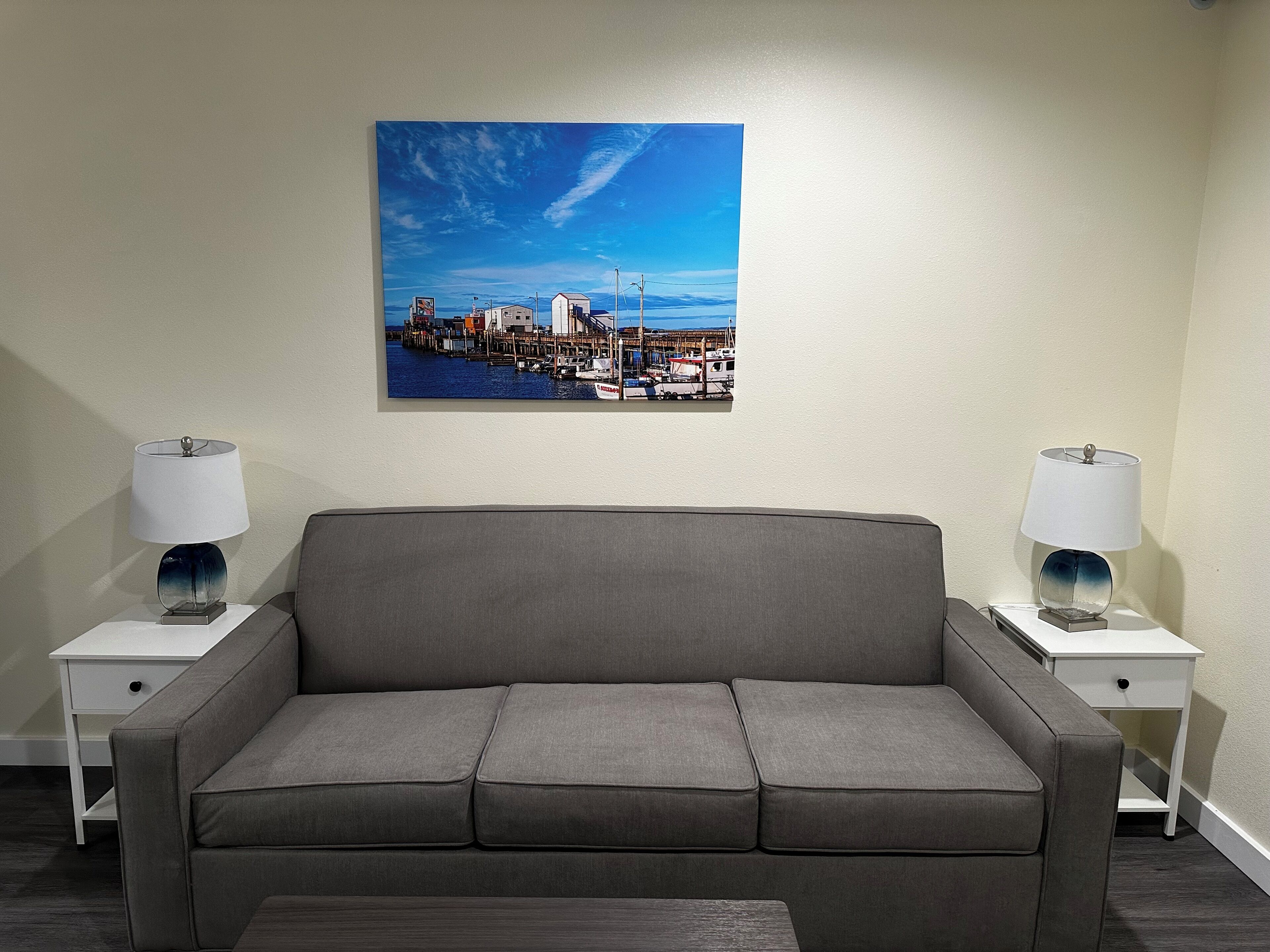 A modern living room with a grey sofa, two bedside tables with lamps, and a cityscape painting on the wall.
