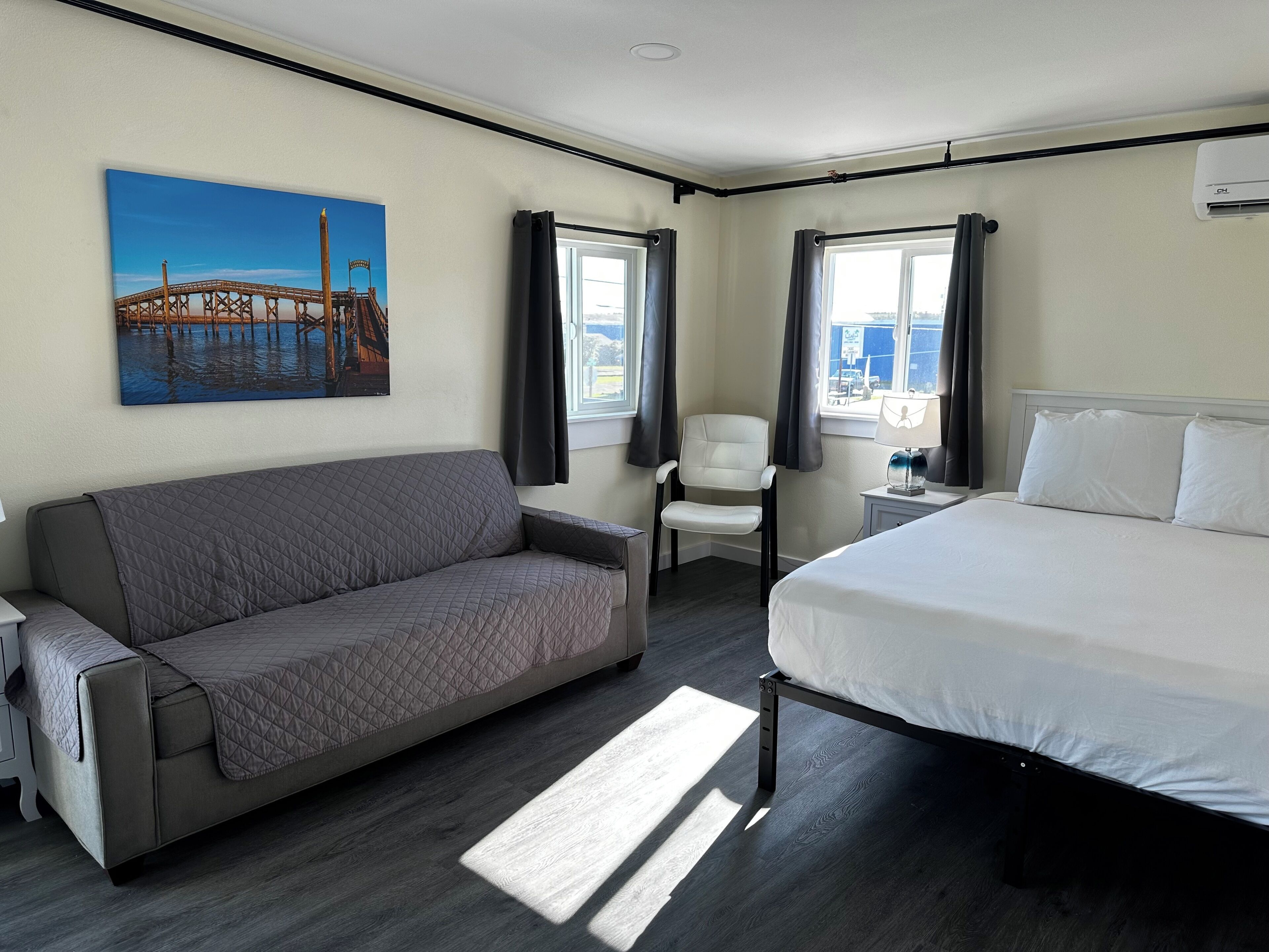 A hotel room with a bed, a sofa, a chair, a window with curtains, and a painting of a bridge.