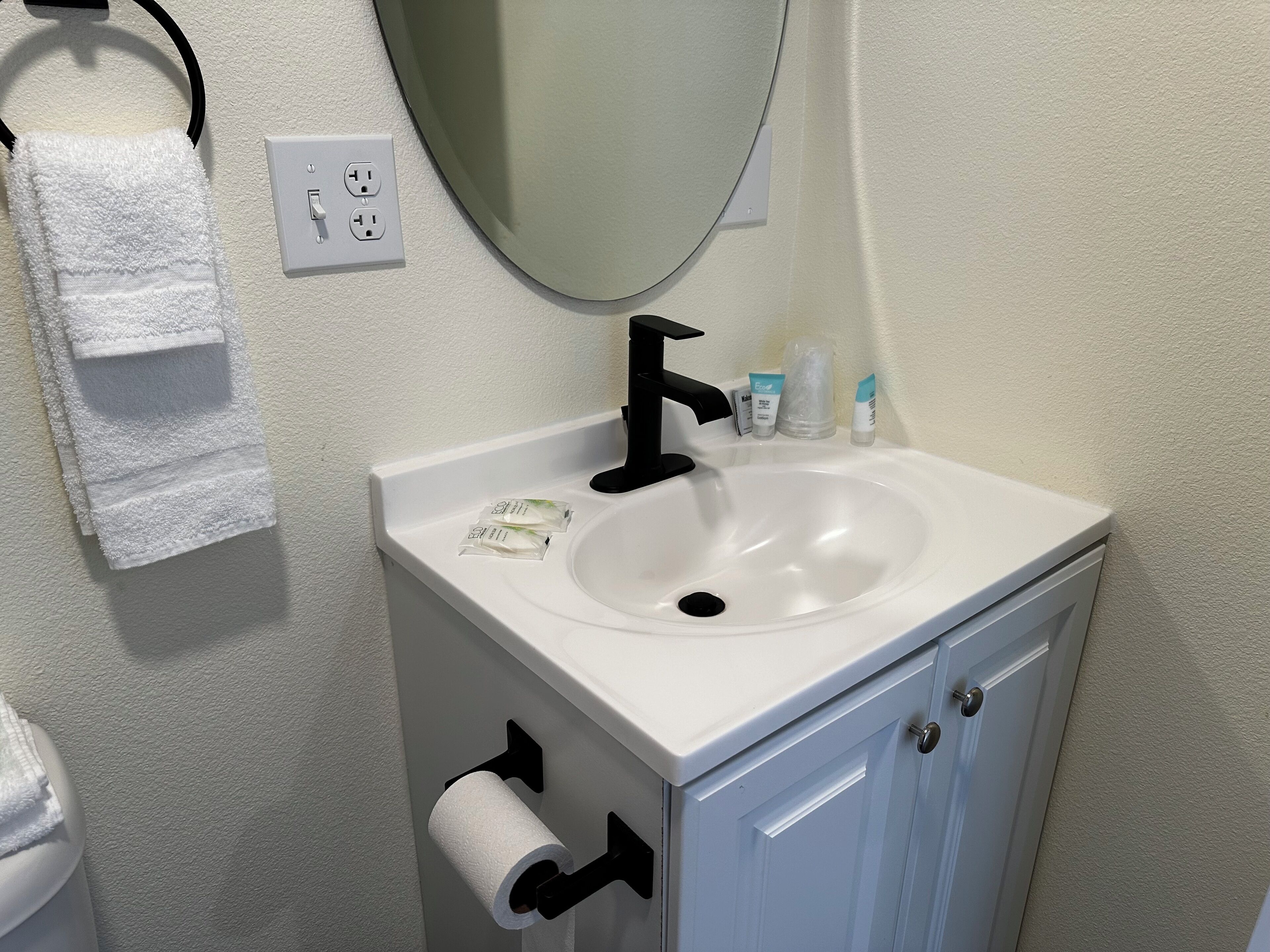 A bathroom with a white sink, black faucet, and a mirror above it. There are toiletries on the sink and a towel rack with a white towel.