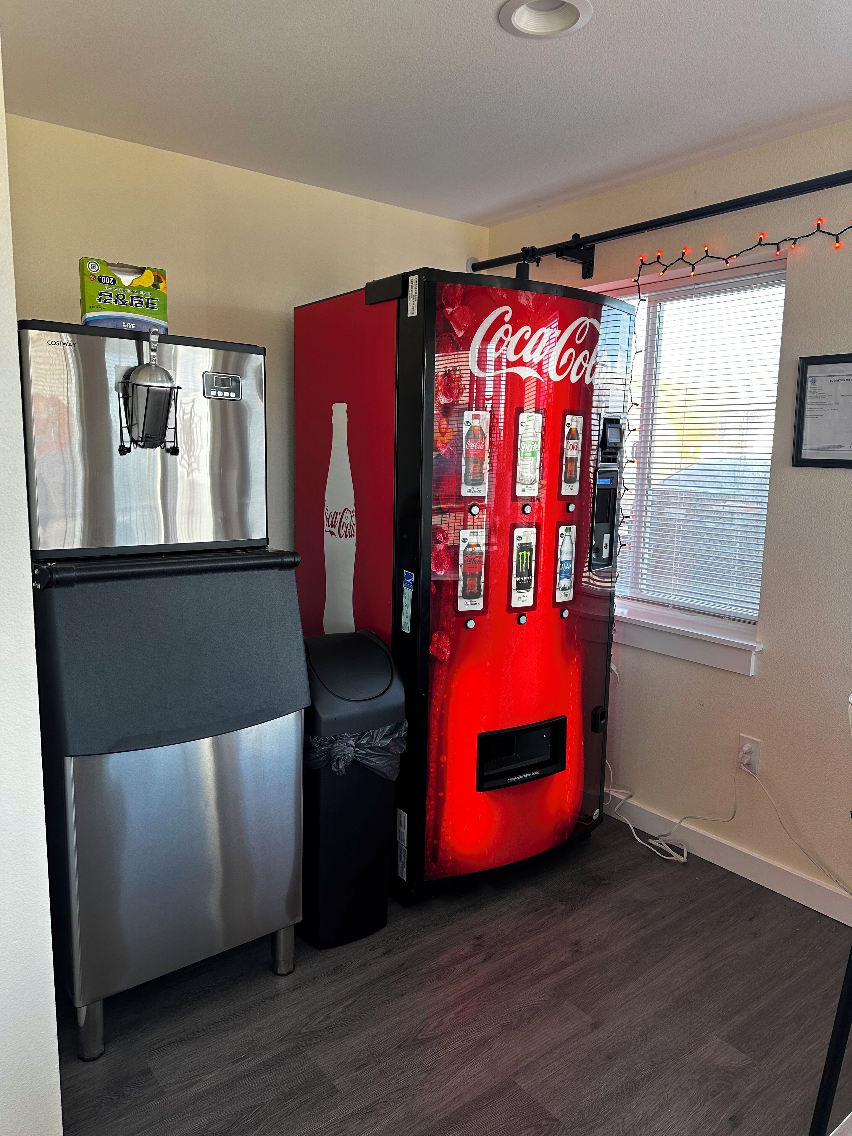 A compact kitchenette with a stainless steel refrigerator, a red Coca-Cola branded beverage dispenser, and a trash bin.