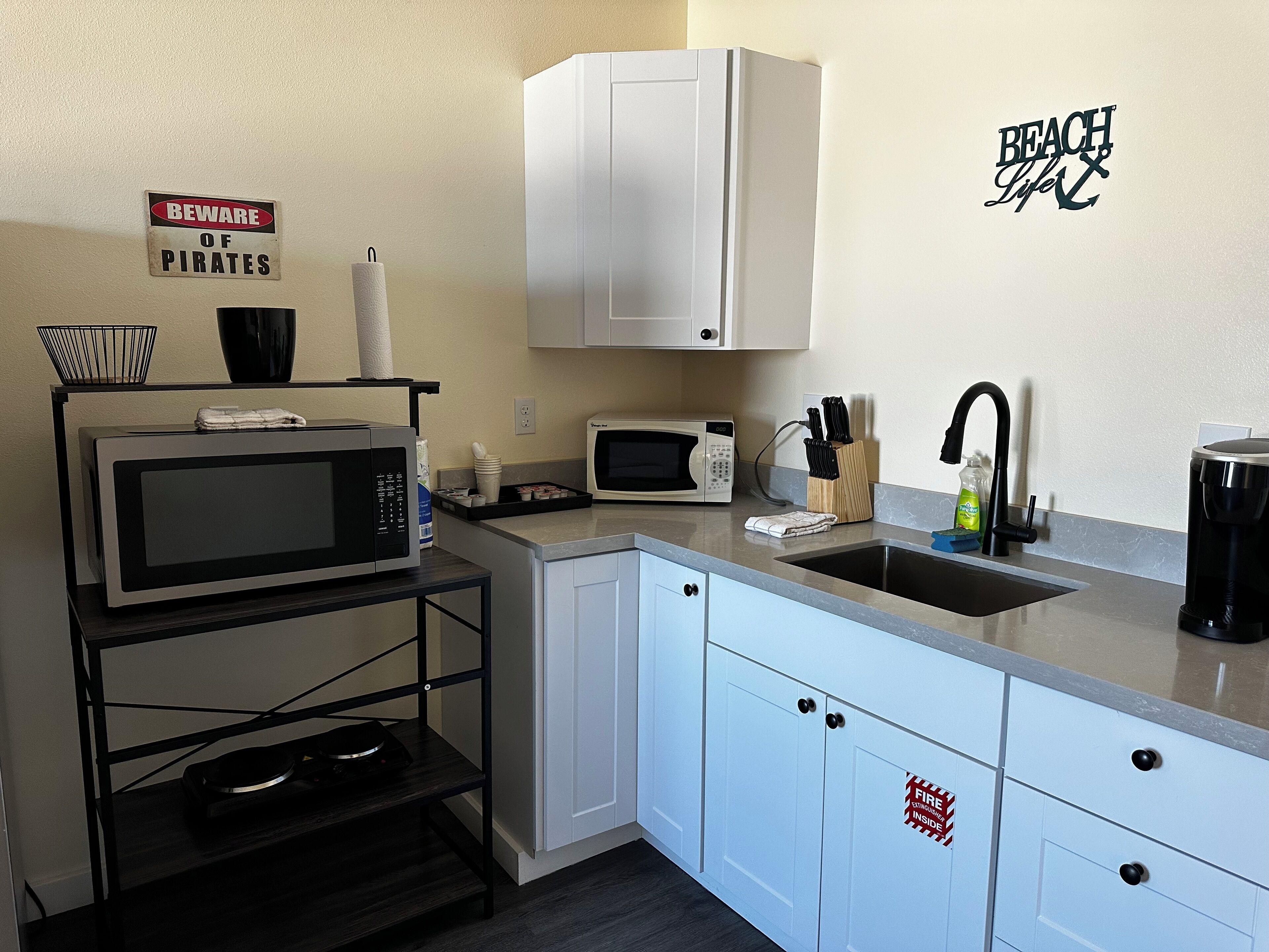 A compact kitchen with white cabinets, a microwave, a toaster, and a sink. A 'Beware of Pirates' sign is visible on the wall.