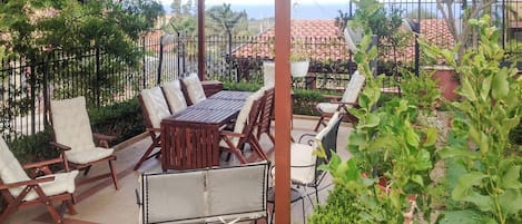 An outdoor patio with a wooden table and chairs, surrounded by a metal fence and greenery.