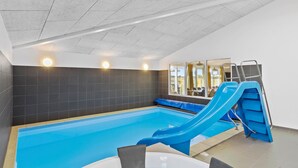 Una piscina interior con un tobogán azul, un techo iluminado por el sol y vistas al exterior a través de amplias ventanas.