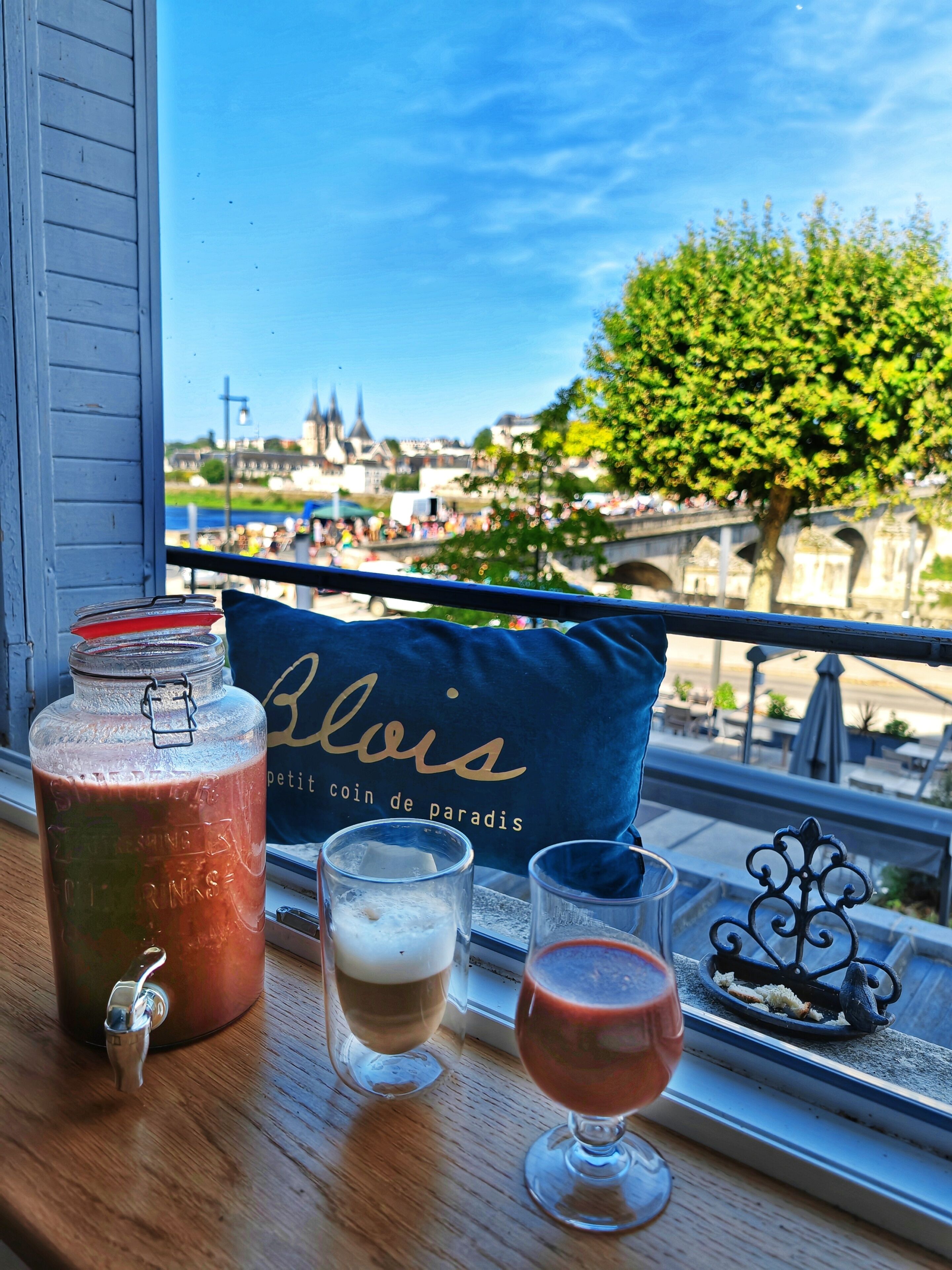 A table with a beverage dispenser, two glasses, and a bottle, with a view of a cityscape and a tree in the background.