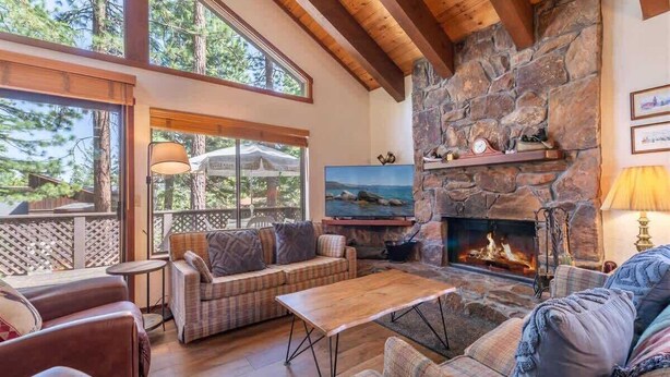 A cozy living room with a stone fireplace, comfortable seating, and a large window overlooking trees. - Viani Cabin (Carnelian Bay)