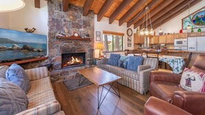 A cozy living room with a stone fireplace, wooden beams, a television, and a dining area with wooden furniture. - Viani Cabin (Carnelian Bay)