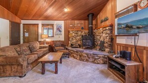 A cozy living room with a stone fireplace, wooden ceiling, and a large window with a view of the sea.