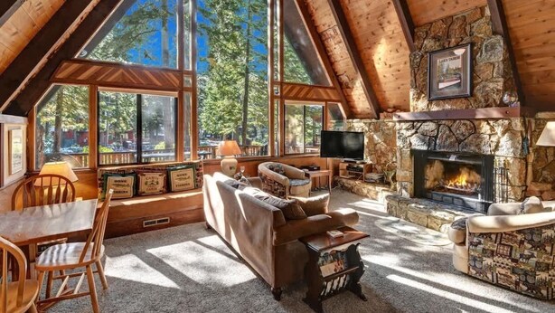 L’intérieur chaleureux d’un chalet avec une cheminée en pierre, des poutres en bois et de grandes fenêtres donnant sur une forêt.
