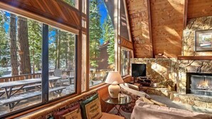 Un intérieur de chalet chaleureux, avec de grandes fenêtres, une cheminée en pierre et une vue sur la forêt.