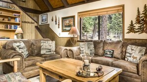 A rustic cabin interior with leather sofas, a wooden coffee table, and a bookshelf.