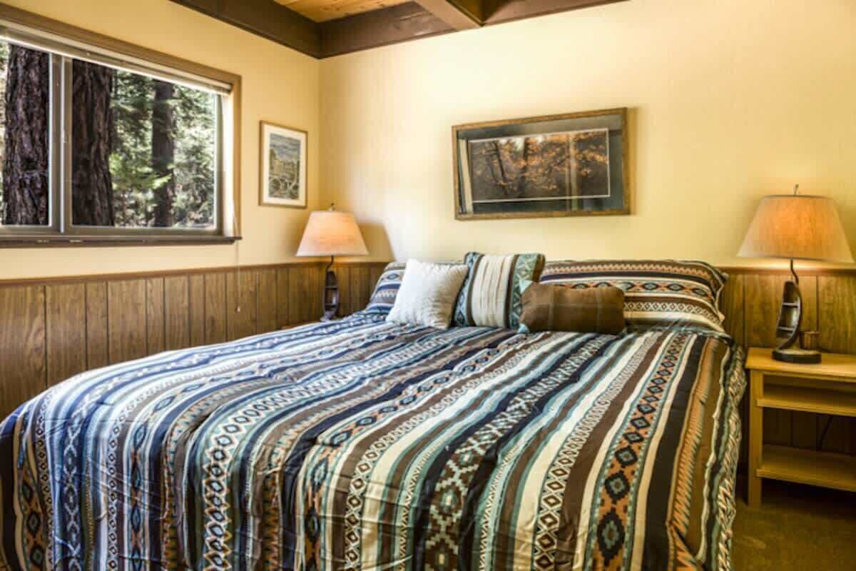 Quarto com uma cama grande, duas luminárias de cabeceira, um quadro na parede e uma janela com vista para árvores.
