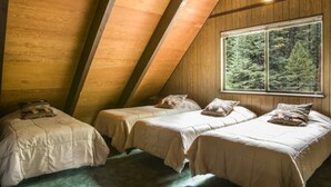 A cozy attic bedroom with three beds, wooden walls, and a large window offering a forest view.