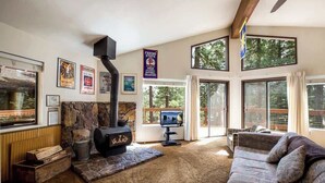 A cozy living room with a stone fireplace, a couch, and large windows offering a view of the outdoors. - Yemoto Cabin (Carnelian Bay)