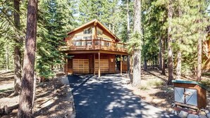 A two-story log cabin with a covered front porch and a garage, surrounded by tall pine trees. - Yemoto Cabin (Carnelian Bay)