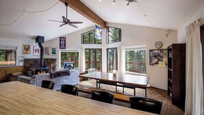 A spacious living room with a large wooden table, comfortable seating, a fireplace, and large windows offering views of the outdoors. - Yemoto Cabin (Carnelian Bay)