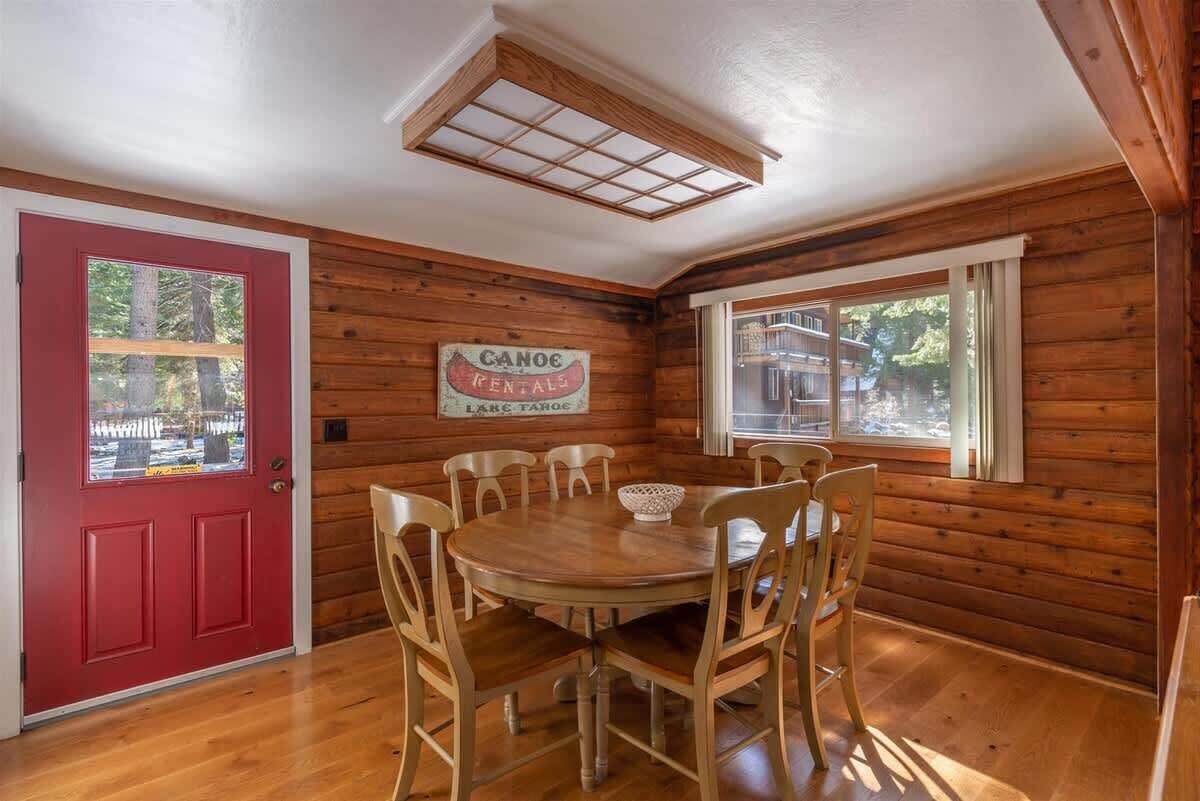 A wooden cabin interior with a red door, a dining table with chairs, and a window.