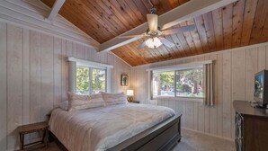 A bedroom with a wooden ceiling, a bed, a nightstand, and a window with a view of trees. - Zoss Cabin (Carnelian Bay)
