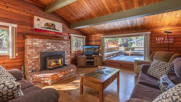 A cozy cabin interior with a brick fireplace, wooden ceiling, and a large window overlooking a deck. - Zoss Cabin (Carnelian Bay)