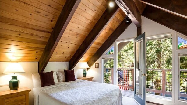 A bedroom with a large bed, wooden ceiling beams, a nightstand, and a window with a view of greenery. - O`Rourke Cabin, 4BD/2BA (Carnelian Bay)