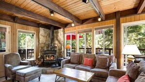A cozy living room with a stone fireplace, wooden beams, and large windows offering a view of the outdoors. - O`Rourke Cabin, 4BD/2BA (Carnelian Bay)