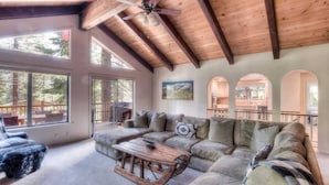 A spacious living room with a large sectional sofa, a wooden coffee table, and a ceiling fan. There are large windows providing natural light and views of the outdoors.
