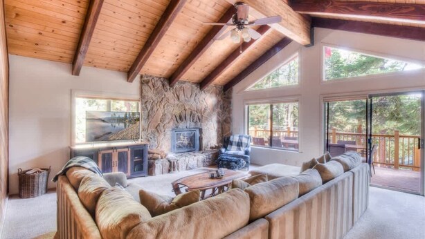 A spacious living room with a stone fireplace, large windows, and a wooden ceiling.