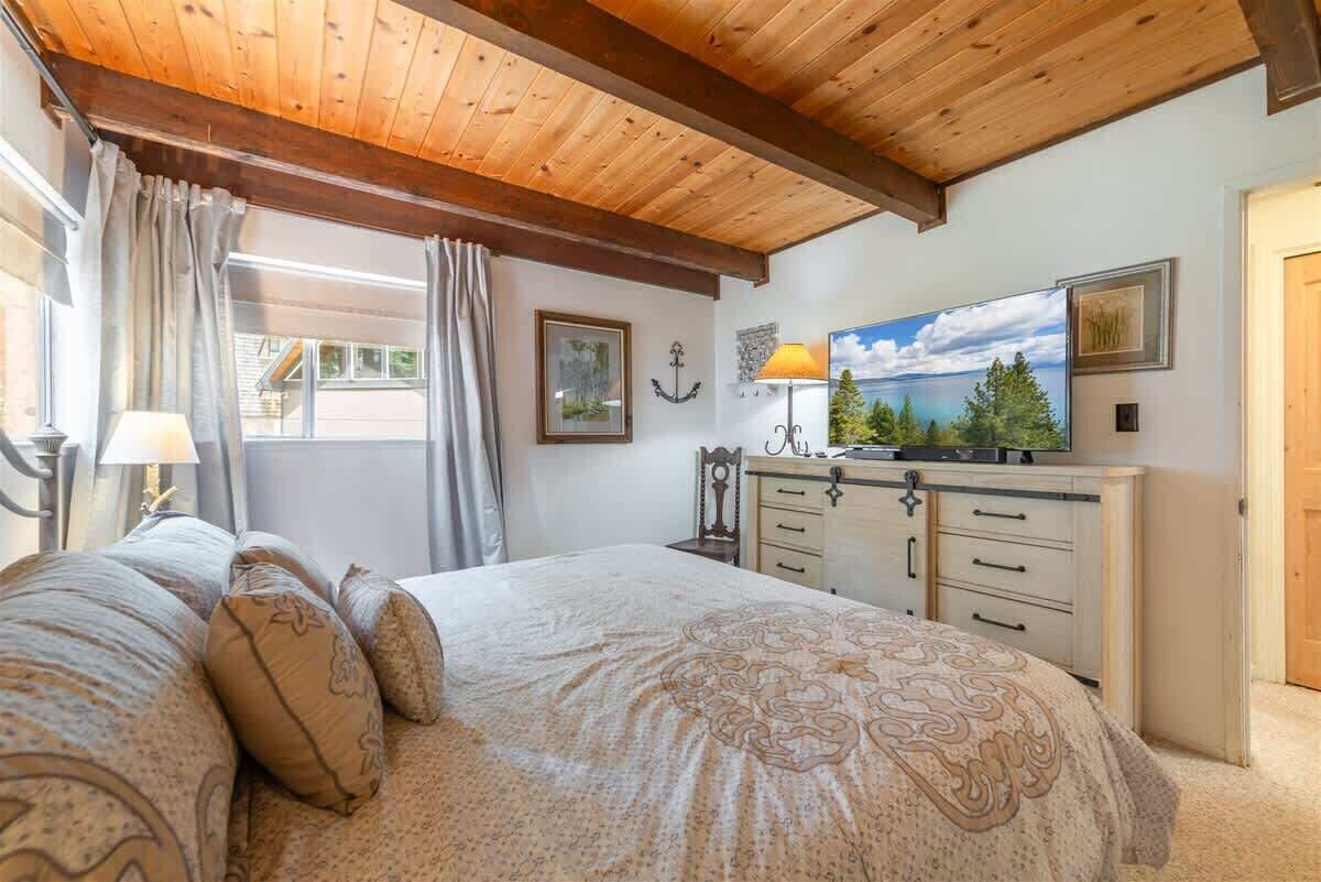 A bedroom with a wooden ceiling, a bed with patterned bedding, a dresser, and a TV on a stand.