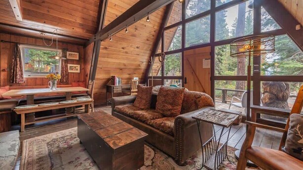 A cozy cabin interior with a wooden dining area, a sofa, and a large window offering a forest view. - Sunshine Cabin (Carnelian Bay)